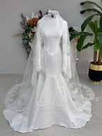 White mermaid satin-look hijab wedding dress with a high neckline, long sleeves, fitted bodice and floor-length train, styled with an intricate floral lace bridal veil. Fatma Sevildi Haute Couture modest bridal design.