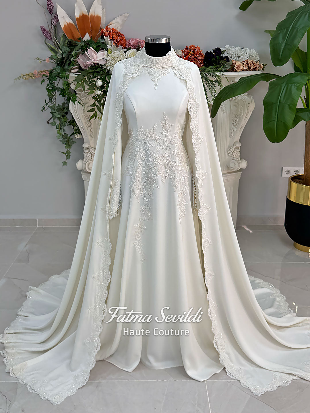 Ivory long sleeve hijab wedding dress with detachable lace-edged cape, floral beaded embroidery, high neckline and floor-length A-line modest bridal silhouette. Designed for Muslim brides seeking an elegant luxury wedding gown with full coverage.