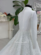 White A-line hijab wedding dress with a high-neck satin bodice, long sleeves, and a beaded botanical lace tulle skirt with a sweeping train. Displayed beside a coordinating lace-trimmed veil.