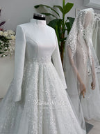 White A-line hijab wedding dress with a high-neck satin bodice, long sleeves, and a beaded botanical lace tulle skirt with a sweeping train. Displayed beside a coordinating lace-trimmed veil.
