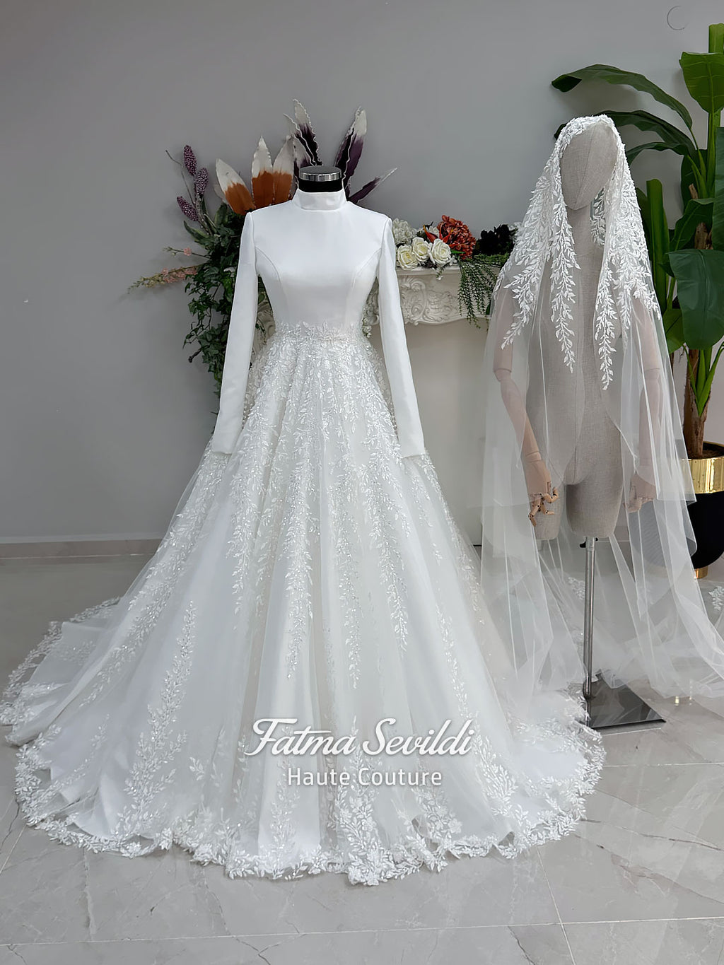 White A-line hijab wedding dress with a high-neck satin bodice, long sleeves, and a beaded botanical lace tulle skirt with a sweeping train. Displayed beside a coordinating lace-trimmed veil.