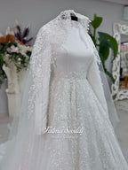 White A-line hijab wedding dress with a high-neck satin bodice, long sleeves, and a beaded botanical lace tulle skirt with a sweeping train. Displayed beside a coordinating lace-trimmed veil.