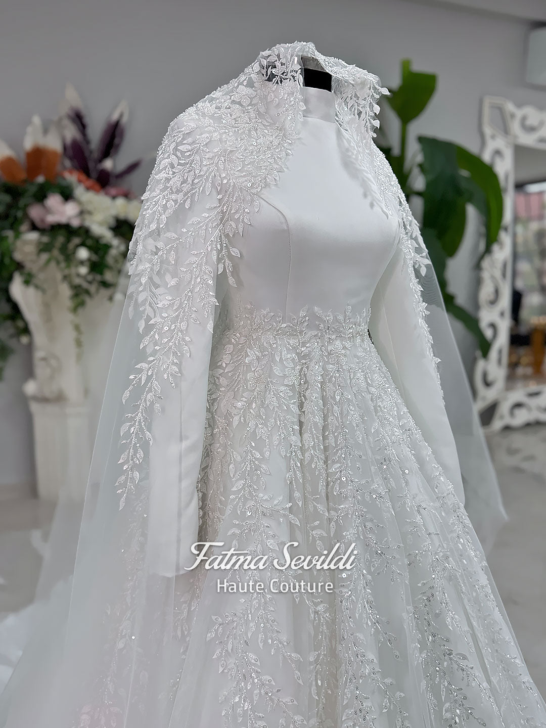 White A-line hijab wedding dress with a high-neck satin bodice, long sleeves, and a beaded botanical lace tulle skirt with a sweeping train. Displayed beside a coordinating lace-trimmed veil.