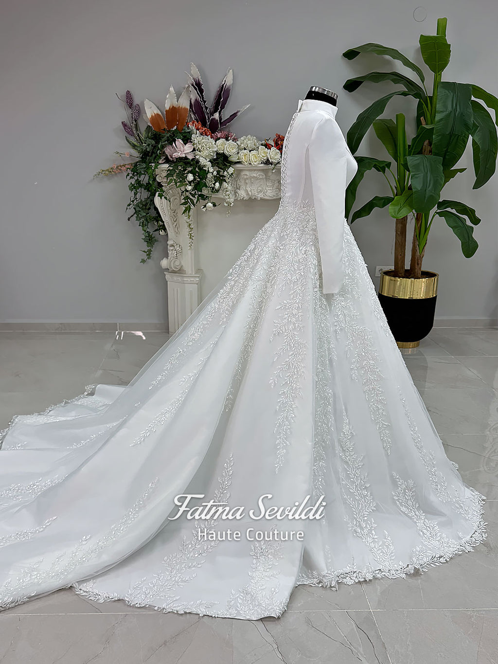 White A-line hijab wedding dress with a high-neck satin bodice, long sleeves, and a beaded botanical lace tulle skirt with a sweeping train. Displayed beside a coordinating lace-trimmed veil.