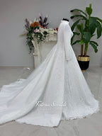 White A-line hijab wedding dress with a high-neck satin bodice, long sleeves, and a beaded botanical lace tulle skirt with a sweeping train. Displayed beside a coordinating lace-trimmed veil.