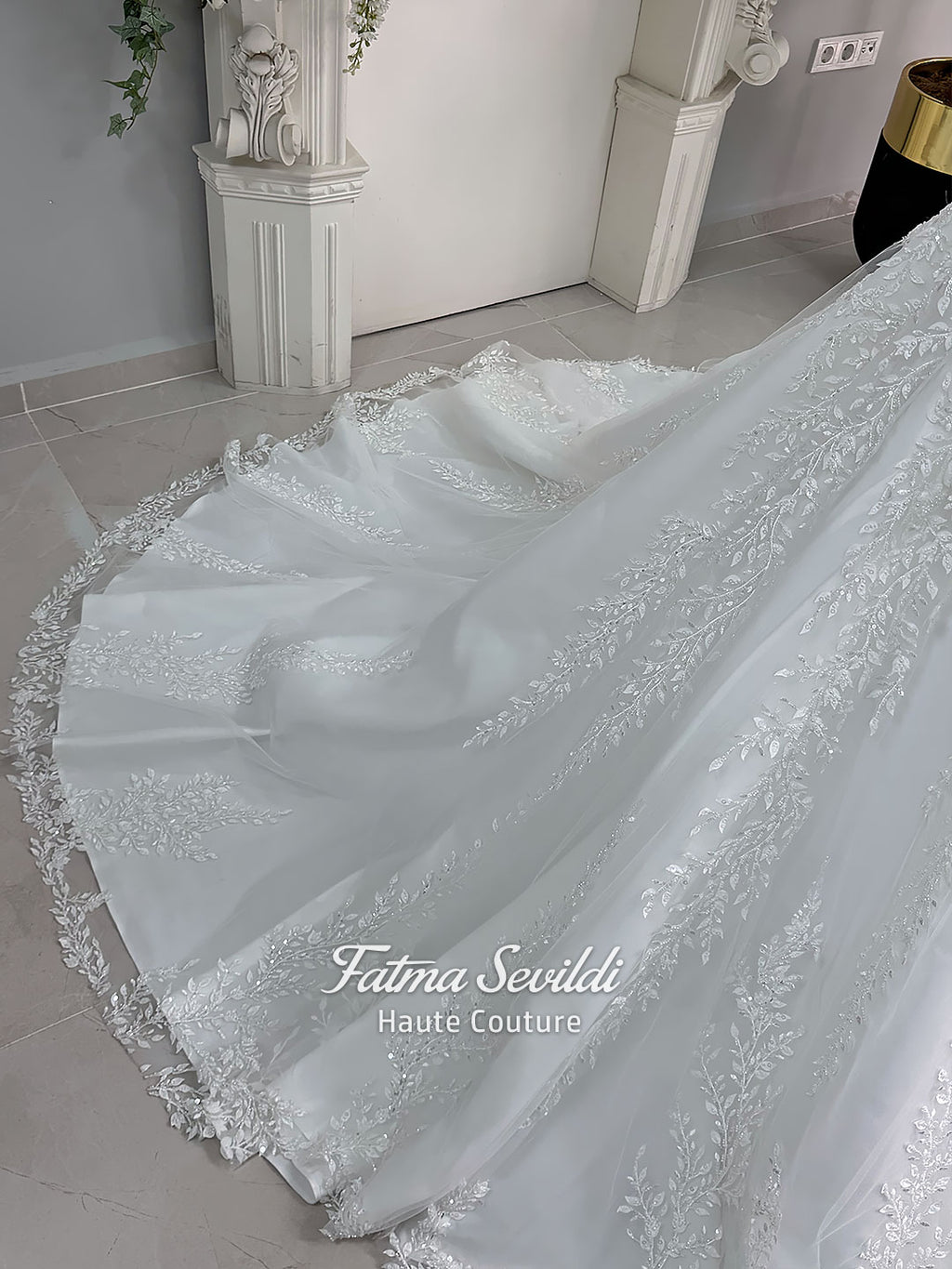 White A-line hijab wedding dress with a high-neck satin bodice, long sleeves, and a beaded botanical lace tulle skirt with a sweeping train. Displayed beside a coordinating lace-trimmed veil.