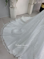 White A-line hijab wedding dress with a high-neck satin bodice, long sleeves, and a beaded botanical lace tulle skirt with a sweeping train. Displayed beside a coordinating lace-trimmed veil.