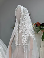White A-line hijab wedding dress with a high-neck satin bodice, long sleeves, and a beaded botanical lace tulle skirt with a sweeping train. Displayed beside a coordinating lace-trimmed veil.