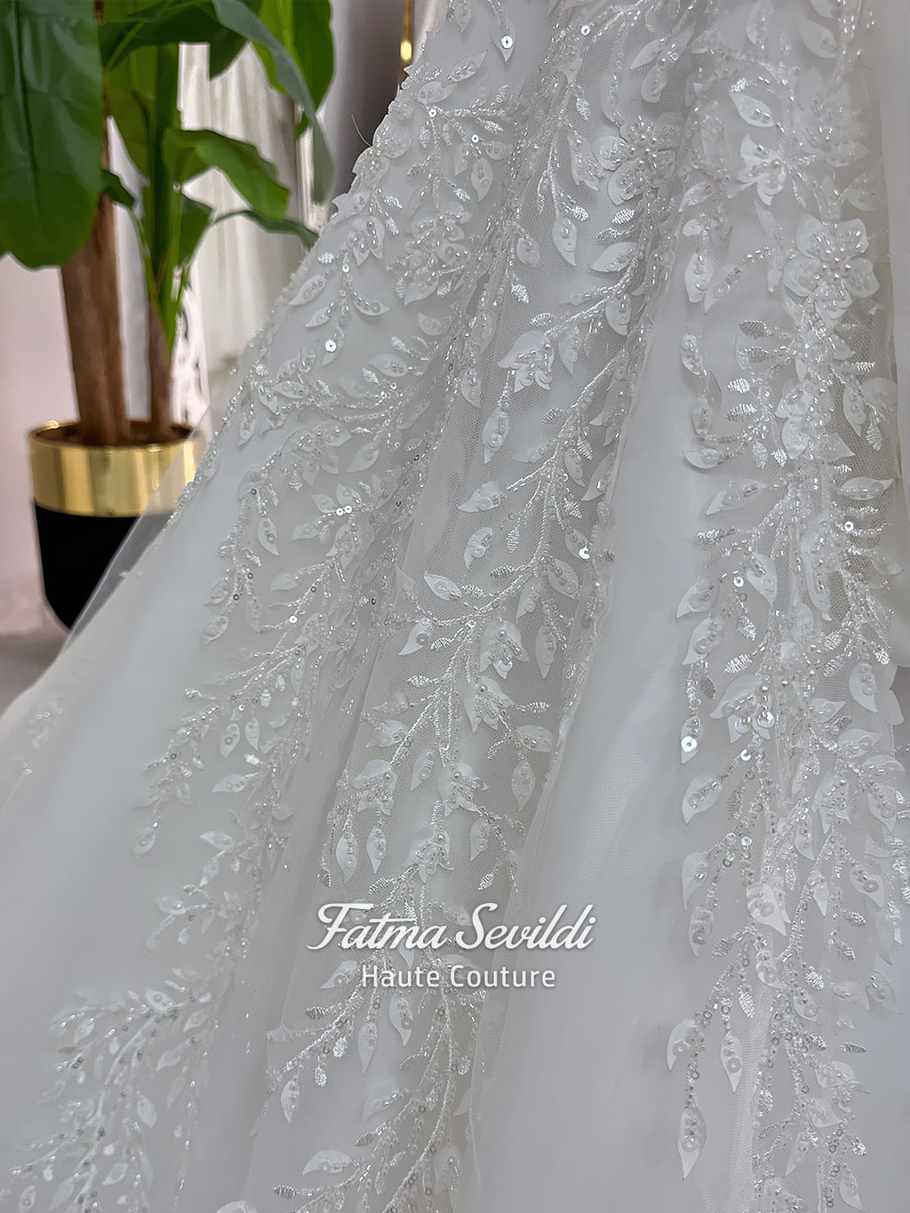 White A-line hijab wedding dress with a high-neck satin bodice, long sleeves, and a beaded botanical lace tulle skirt with a sweeping train. Displayed beside a coordinating lace-trimmed veil.