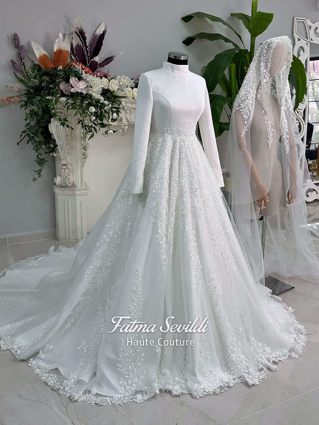 White A-line hijab wedding dress with a high-neck satin bodice, long sleeves, and a beaded botanical lace tulle skirt with a sweeping train. Displayed beside a coordinating lace-trimmed veil.
