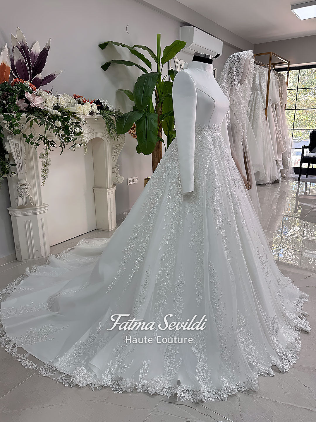 White A-line hijab wedding dress with a high-neck satin bodice, long sleeves, and a beaded botanical lace tulle skirt with a sweeping train. Displayed beside a coordinating lace-trimmed veil.