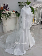 White mermaid satin-look hijab wedding dress with a high neckline, long sleeves, fitted bodice and floor-length train, styled with an intricate floral lace bridal veil. Fatma Sevildi Haute Couture modest bridal design.