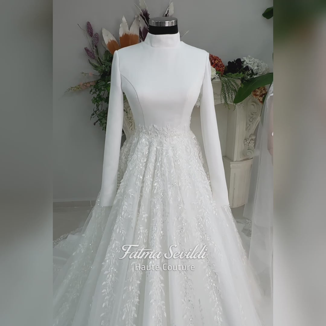 White A-line hijab wedding dress with a high-neck satin bodice, long sleeves, and a beaded botanical lace tulle skirt with a sweeping train. Displayed beside a coordinating lace-trimmed veil.